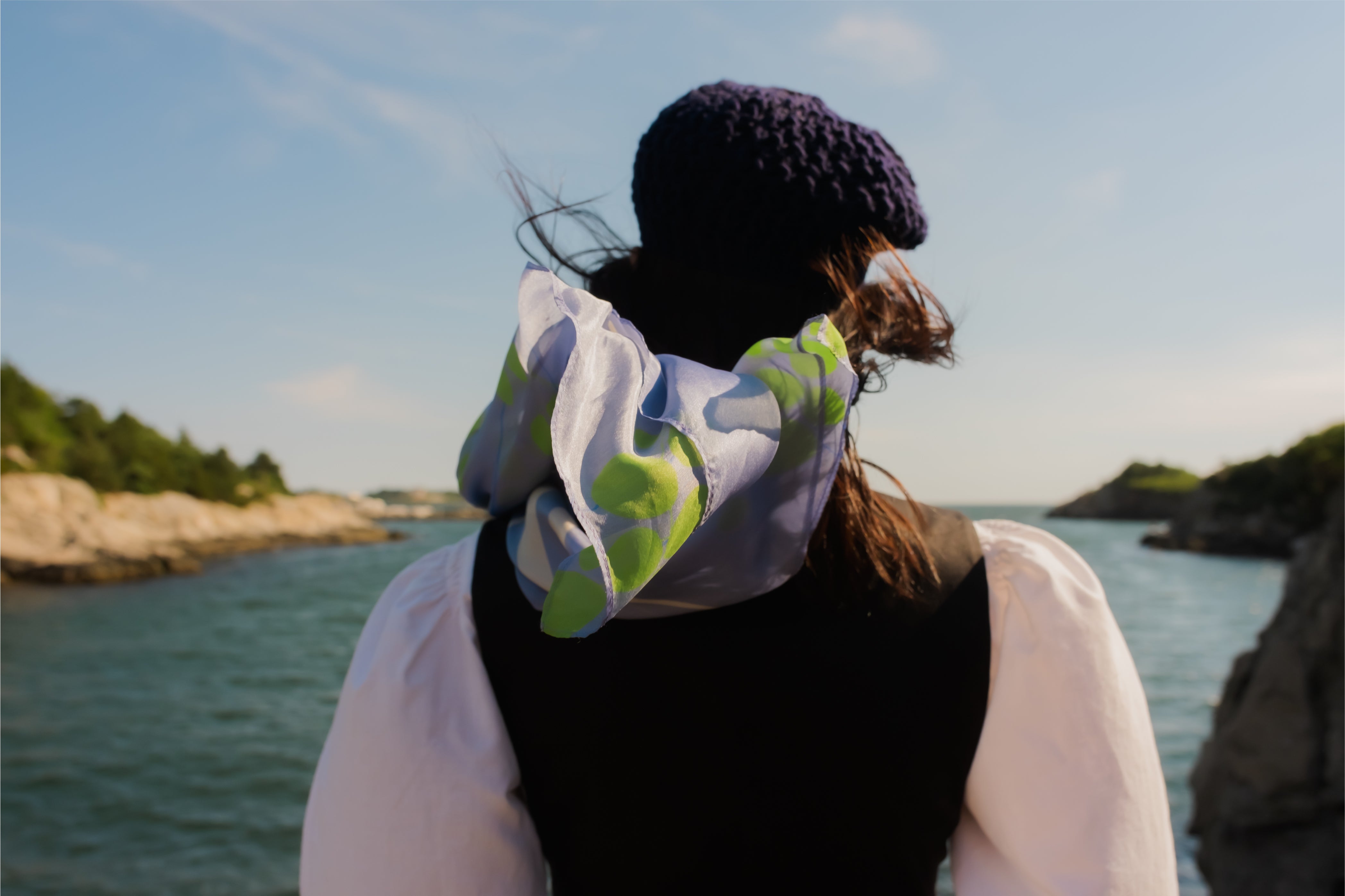 model looks at the ocean and blue, green silk scarf blows behind their shoulders