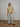 A model stands in a studio wearing a short-sleeve crewneck t-shirt with a geometric pattern in olive green, beige, and taupe, paired with wide-leg light-wash jeans and brown leather loafers.