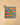 A square fabric pot holder with a colorful striped pattern of orange, teal, blue, and olive green diagonal bands, finished with a brown canvas border and a carrying loop, shown on a light wood background.