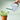 A hand holding a glass watering can pouring water onto a plant on a green background. 
