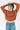 A woman wearing a cropped, ribbed-knit orange sweater and blue jeans, posing with her hands behind her head against a plain white background.