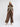 A model stands against a light gray studio background wearing a cream knit beanie, a gray turtleneck, a brown vest with cream faux-fur trim and embroidered details, wide-leg brown plaid trousers, and beige ankle boots.