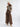 A model stands against a light gray studio background wearing a cream knit beanie, a gray turtleneck, a brown patterned sleeveless vest with cream faux-fur trim, wide-leg brown plaid trousers, and beige pointed-toe boots.