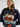 A woman wearing a navy blue knit sweater with a colorful embroidered sailboat and sea-themed design, paired with a beige knit bonnet and black skirt, against a light gray studio background.