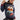 A woman wearing a navy blue knit sweater with a colorful embroidered sailboat and sea-themed design, paired with a beige knit bonnet and black skirt, against a light gray studio background.