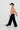 A model wearing a red and cream checkered cropped cardigan, black wide-leg trousers, a black wide-brim hat, and black loafers, walking against a plain white studio background.