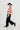 A model is shown in profile wearing a cropped red and cream check knit cardigan, dark wide-leg velvet trousers, a black wide-brim hat, and dark loafers, set against a plain white studio background.