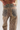 Close-up back view of a person wearing patterned jeans with a brown leather patch on the waistband that reads Tacodhe. The jeans have a mottled brown and gray base with cream speckled spots and dark gray pocket panels.