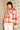 A model wearing a stylish knit cardigan with a red and cream checkered pattern, a matching scarf, a dark brown beanie, and white pants, posed against a vintage floral wallpaper background.