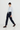 A model walks in profile wearing a light blue long-sleeve tie-neck blouse, dark navy wide-leg jeans, brown leather boots, and a dark brown knit beanie against a clean white studio background.