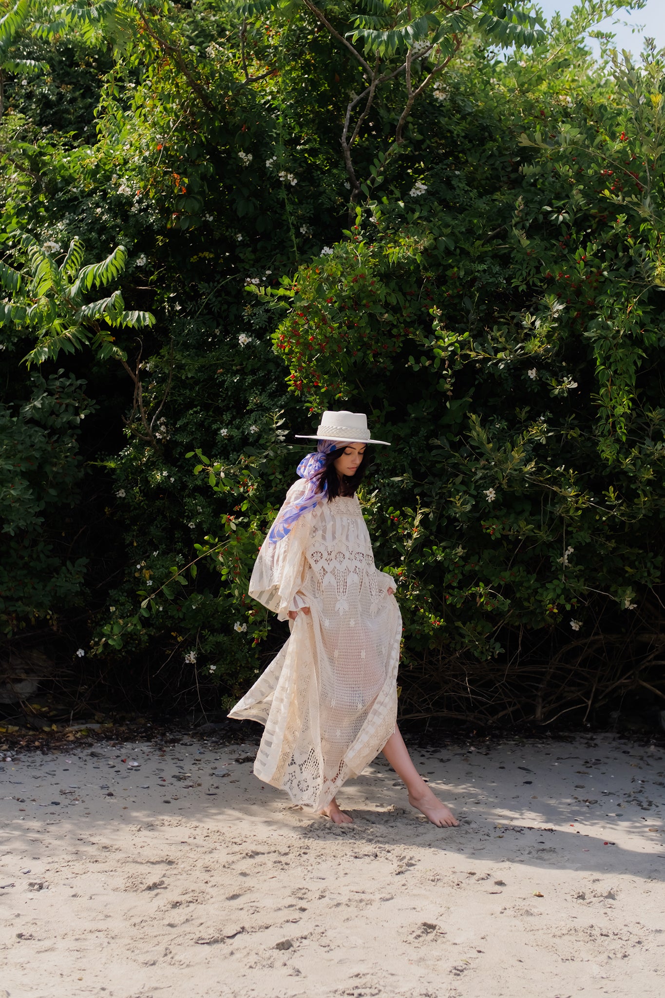 model stands barefoot on a sandy beach in a white chaftan with periwinkle silk scarf