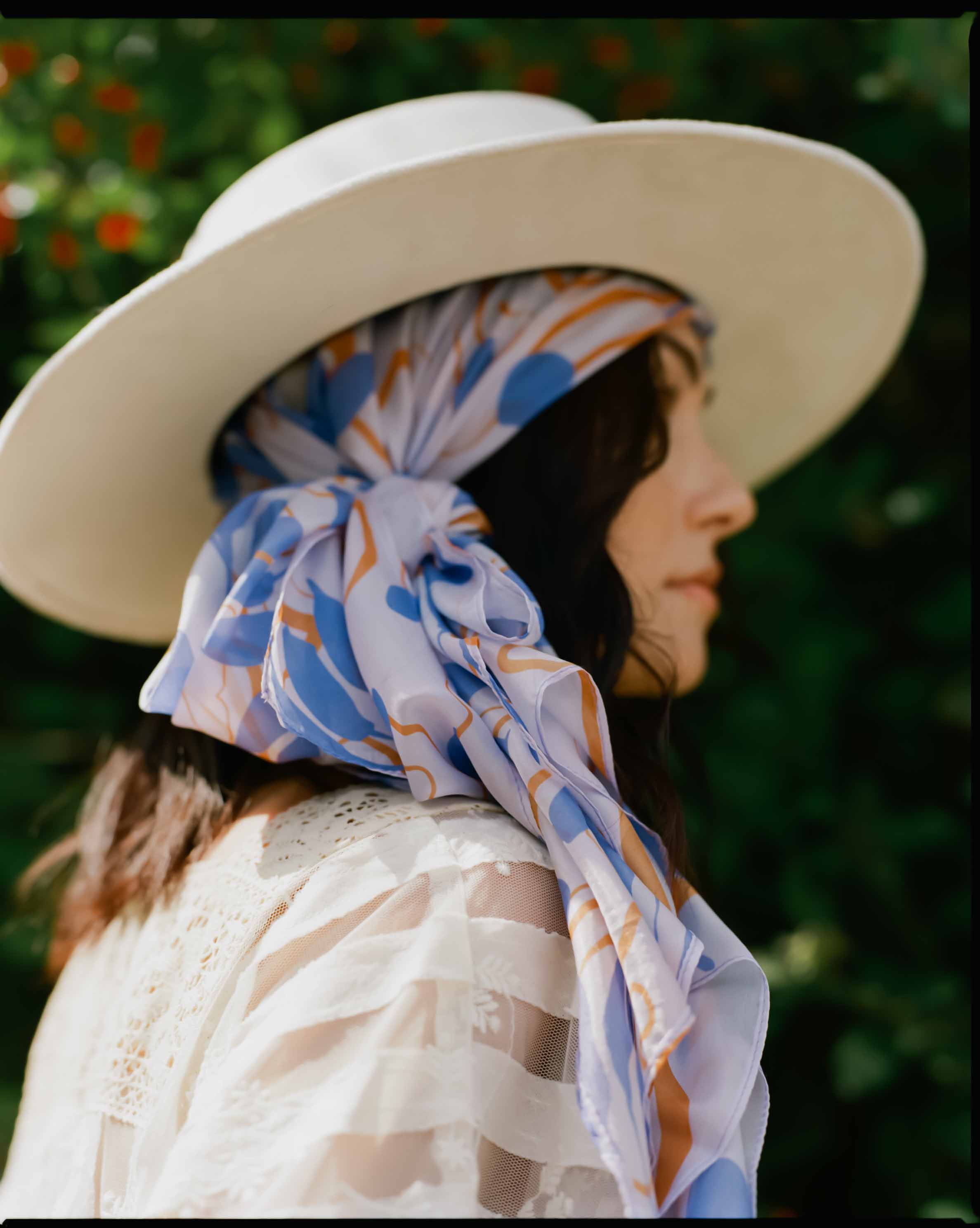 Periwinkle silk scarf in Water Lily Print head wrapped on model and styled under a white flat-brimmed fedora 
