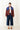 A woman stands on a white background wearing dark blue jeans, brown shoes, and a white shirt with a burgundy, cobalt blue, and navy puffer jacket with cream piping. 