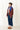 A woman stands on a white background wearing dark blue jeans, brown shoes, and a white shirt with a burgundy, cobalt blue, and navy puffer jacket with cream piping. 